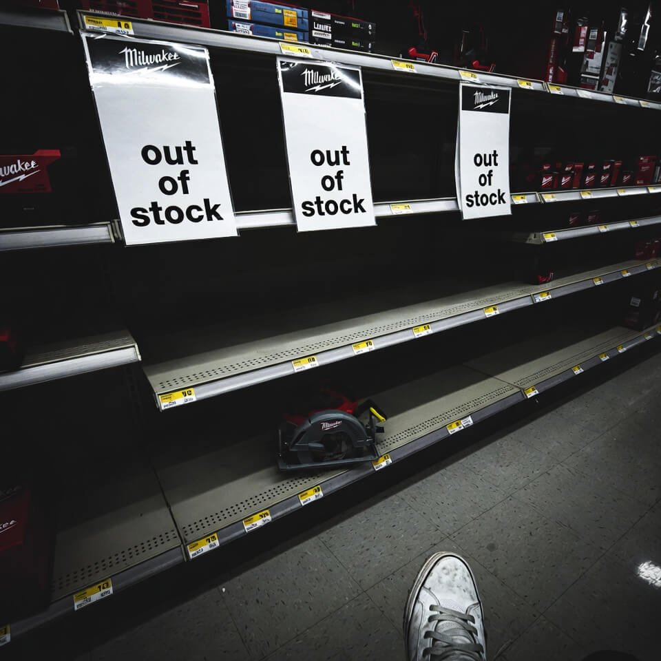 Empty Lowe’s shelf with out-of-stock tags on Milwaukee tools, one lonely circular saw from Vietnam, dirty sneaker in corner.
