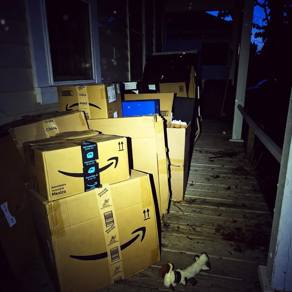 Pile of Amazon packages on porch at dusk, Mexico and China labels, chewed dog toy nearby, yellow porch light.