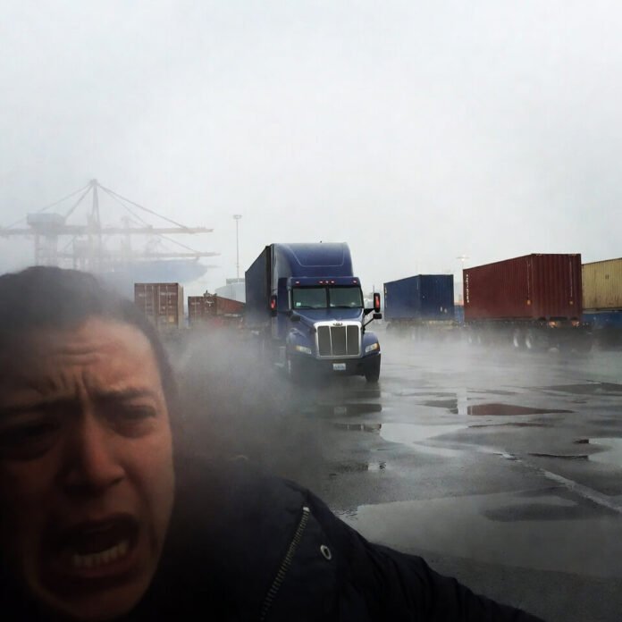 Stressed man checking phone at foggy port with truck and containers.