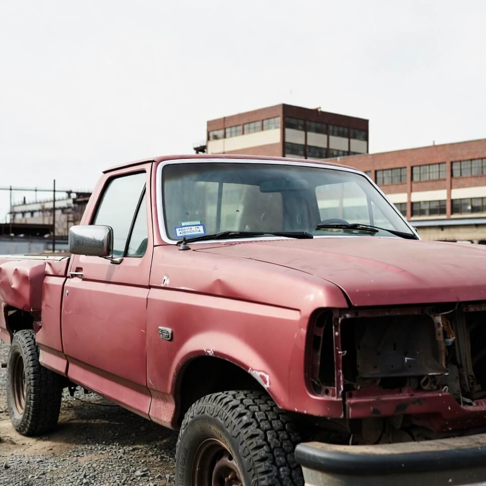 Low-angle view of beat-up old Ford F-150 with peeling "Made in America" sticker outside steel mill.